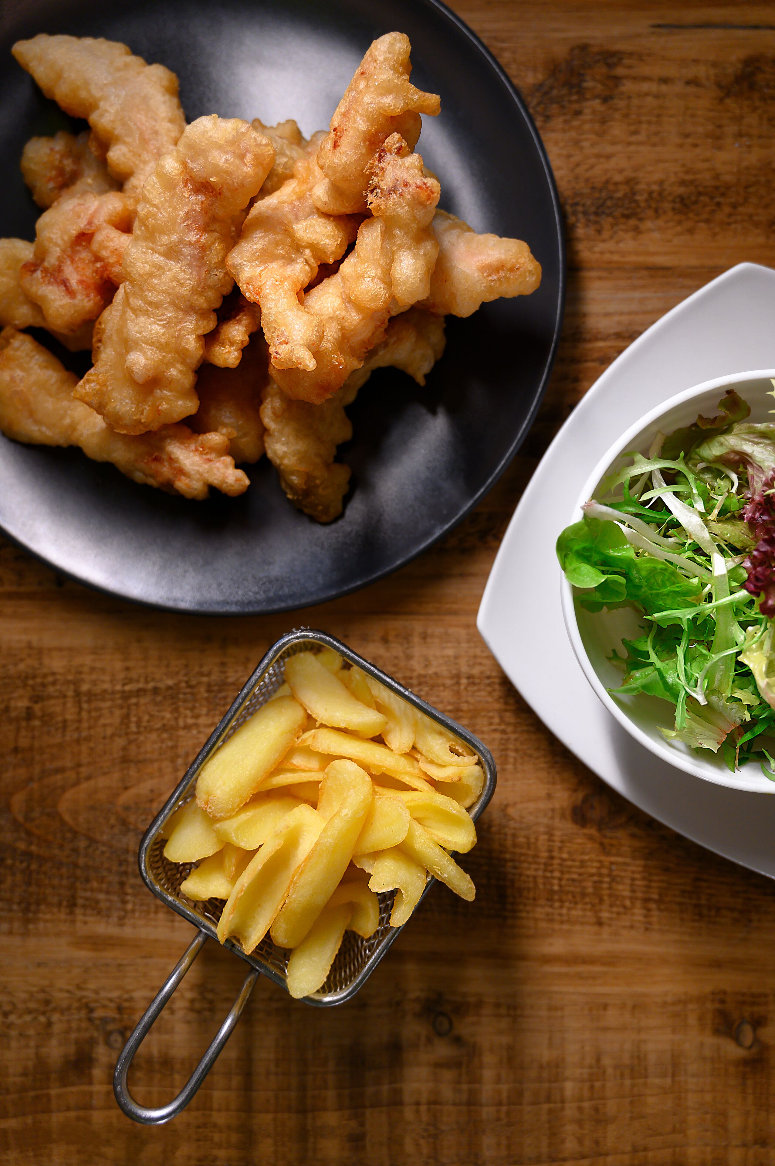 A plate of fried food, a bowl of salad, and a colander with fries on a wooden table.