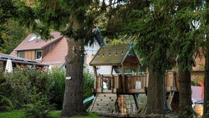 A wooden treehouse with a staircase, surrounded by trees and a house in the background. - Domaine du Hirtz (Wattwiller)