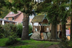 Une cabane en bois perchée dans un arbre, accessible par un escalier, entourée d’arbres, avec une maison en arrière-plan.