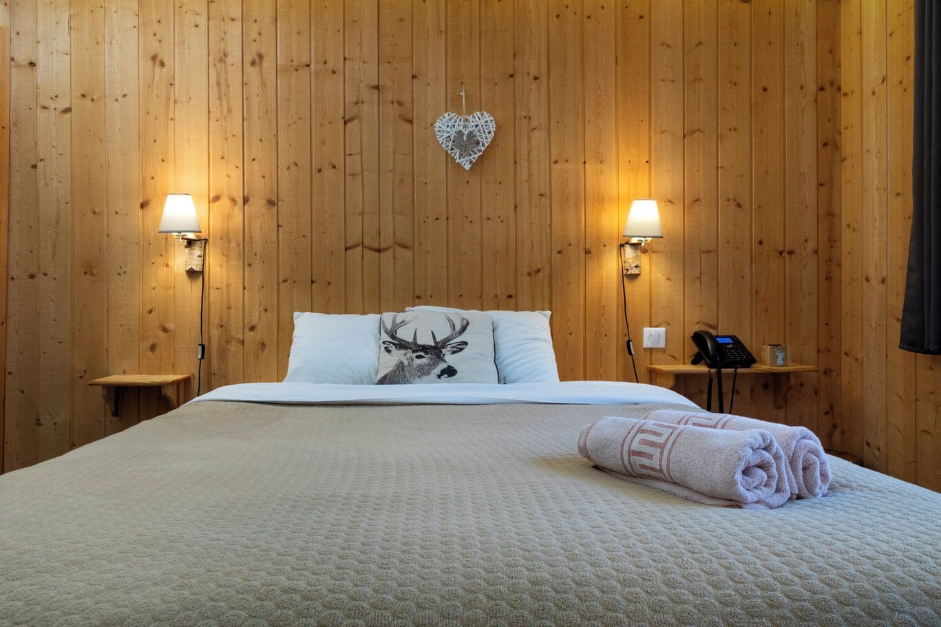 A wooden-paneled room with a bed, two bedside lamps, a heart-shaped decoration, and a phone on a small table.