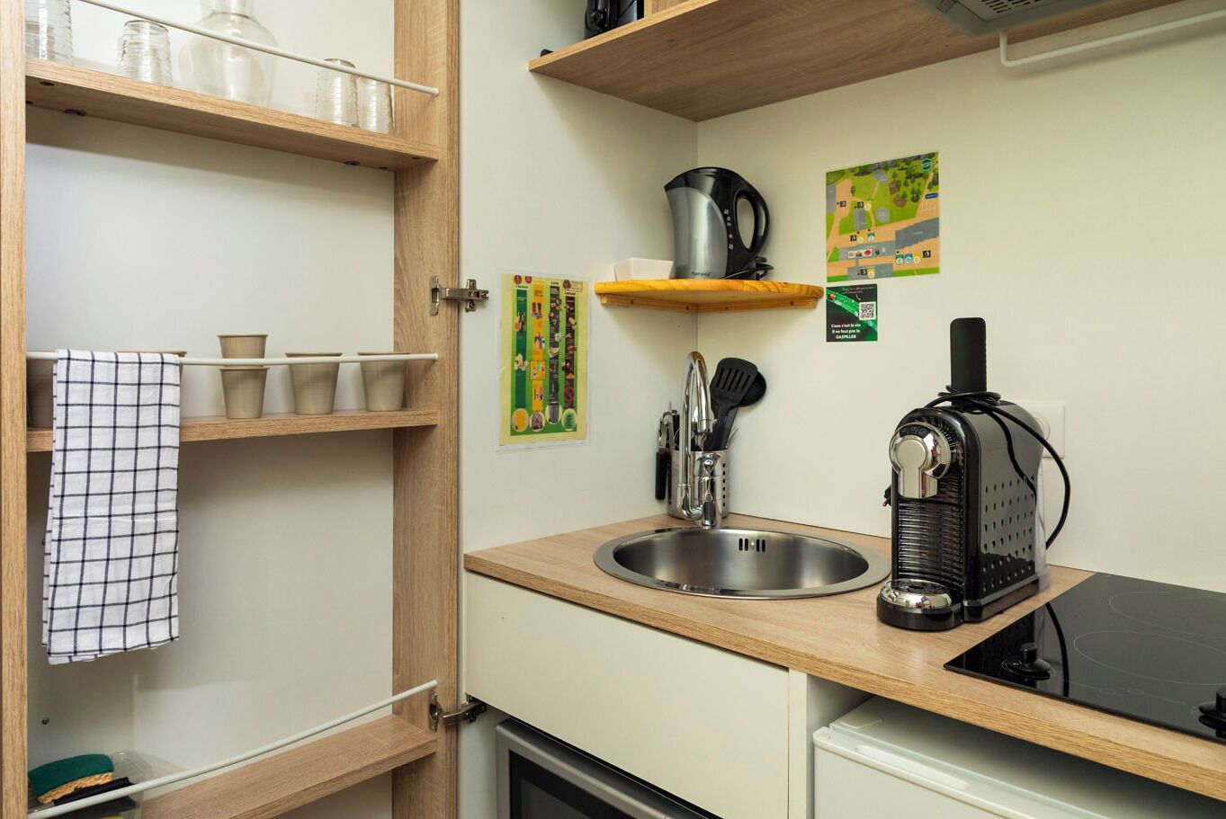 Eine kleine Küche mit Spüle, Wasserkocher und Kaffeemaschine. Regale mit Tassen und eine Karte an der Wand.