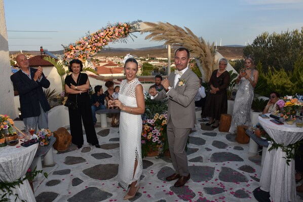 A wedding ceremony with guests seated around tables, a stone floor, and a view of the landscape in the background.