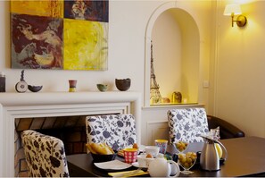 A dining area with a fireplace, floral-patterned chairs, a table set with a basket of bread, cups, and a teapot. - La Villa Paris (Paris)