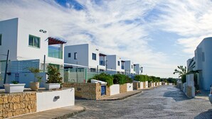 Une rangée de bâtiments modernes aux façades blanches, dotés de murs en pierre et agrémentés de plantes en pot, une rue pavée et un ciel dégagé.