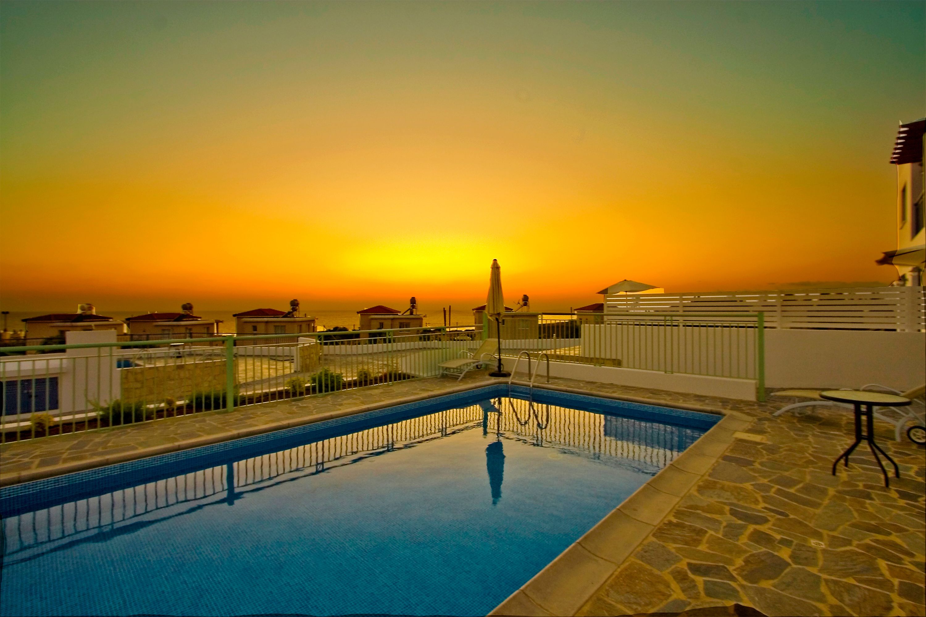 A swimming pool with a stone deck and a view of the sunset over a residential area.