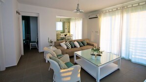Un salon avec un canapé, des fauteuils et une table basse en verre. On aperçoit une cuisine au fond.