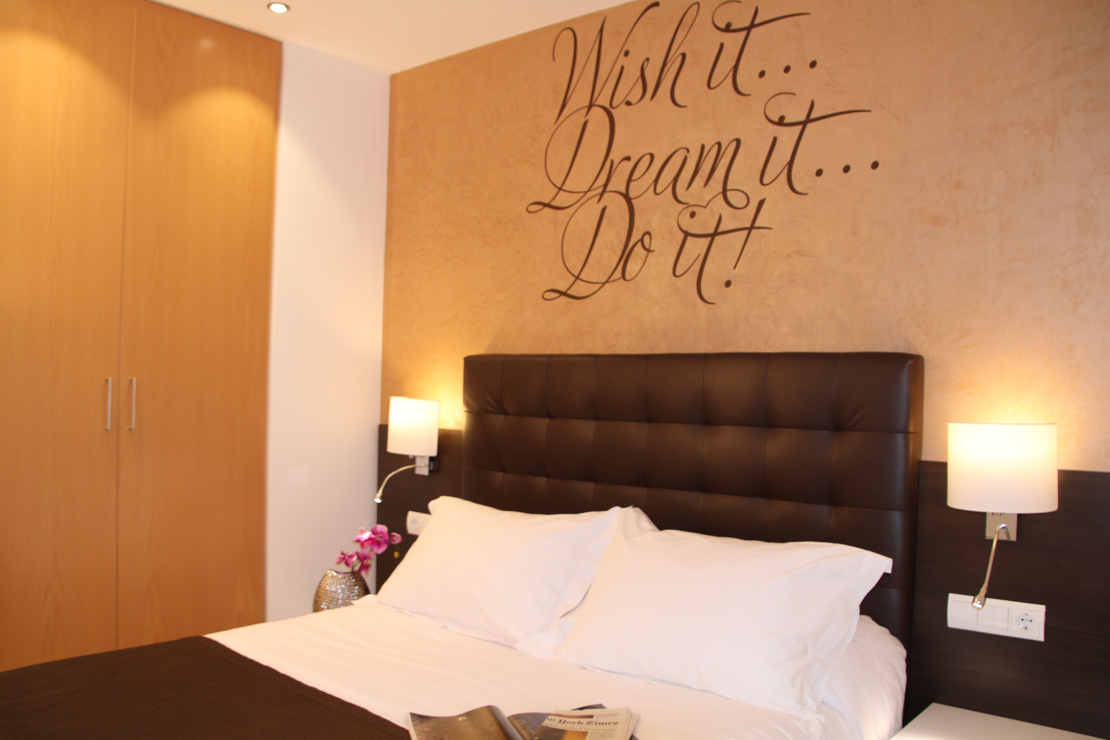 A neatly made bed with white linens, a dark headboard, and a motivational wall quote.