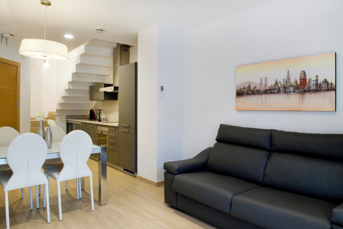 A modern living room with a black sofa, a dining table with white chairs, and a kitchen area with a staircase.