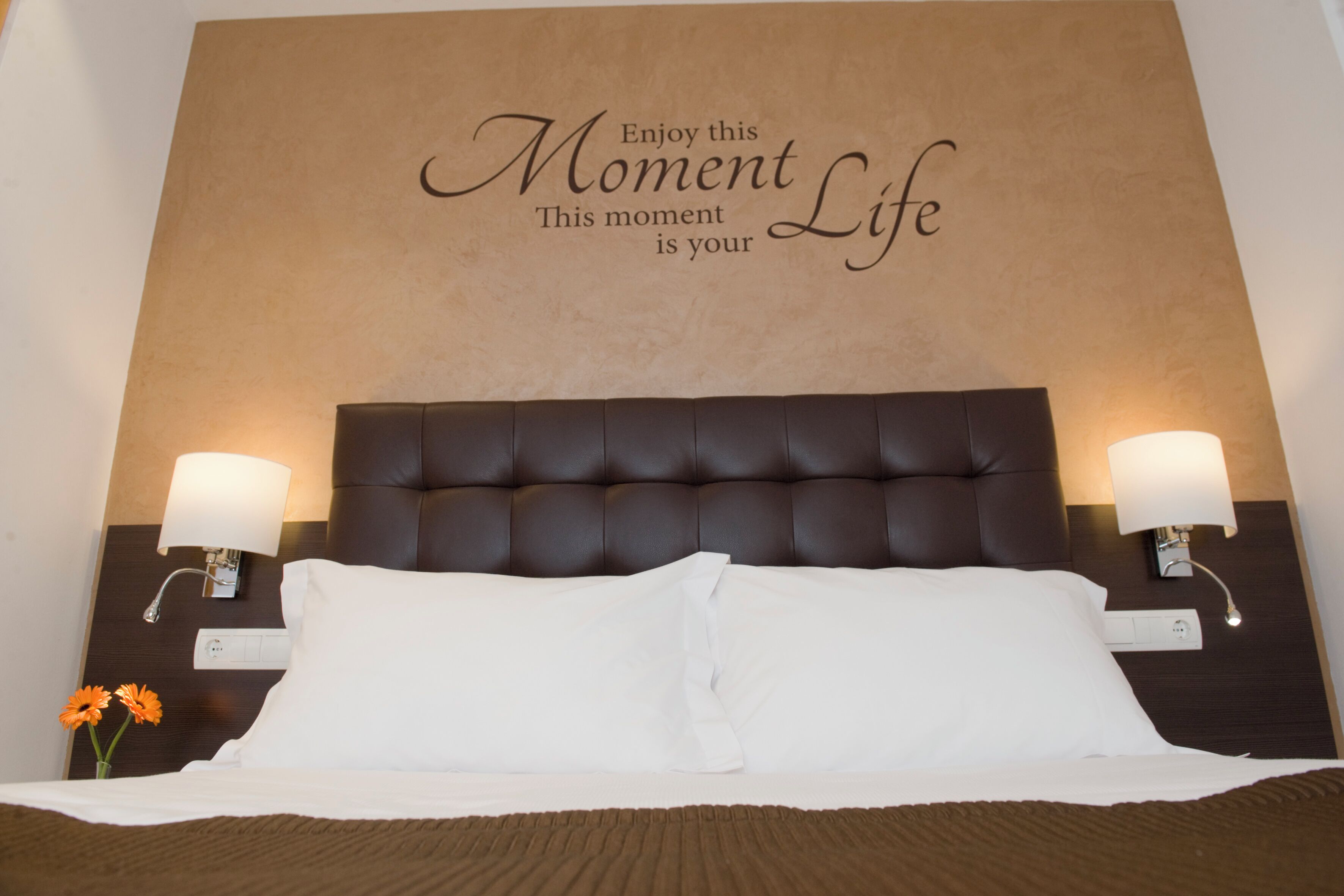 Quarto de hotel com uma cama grande, duas luminárias de cabeceira e uma frase motivacional na parede.