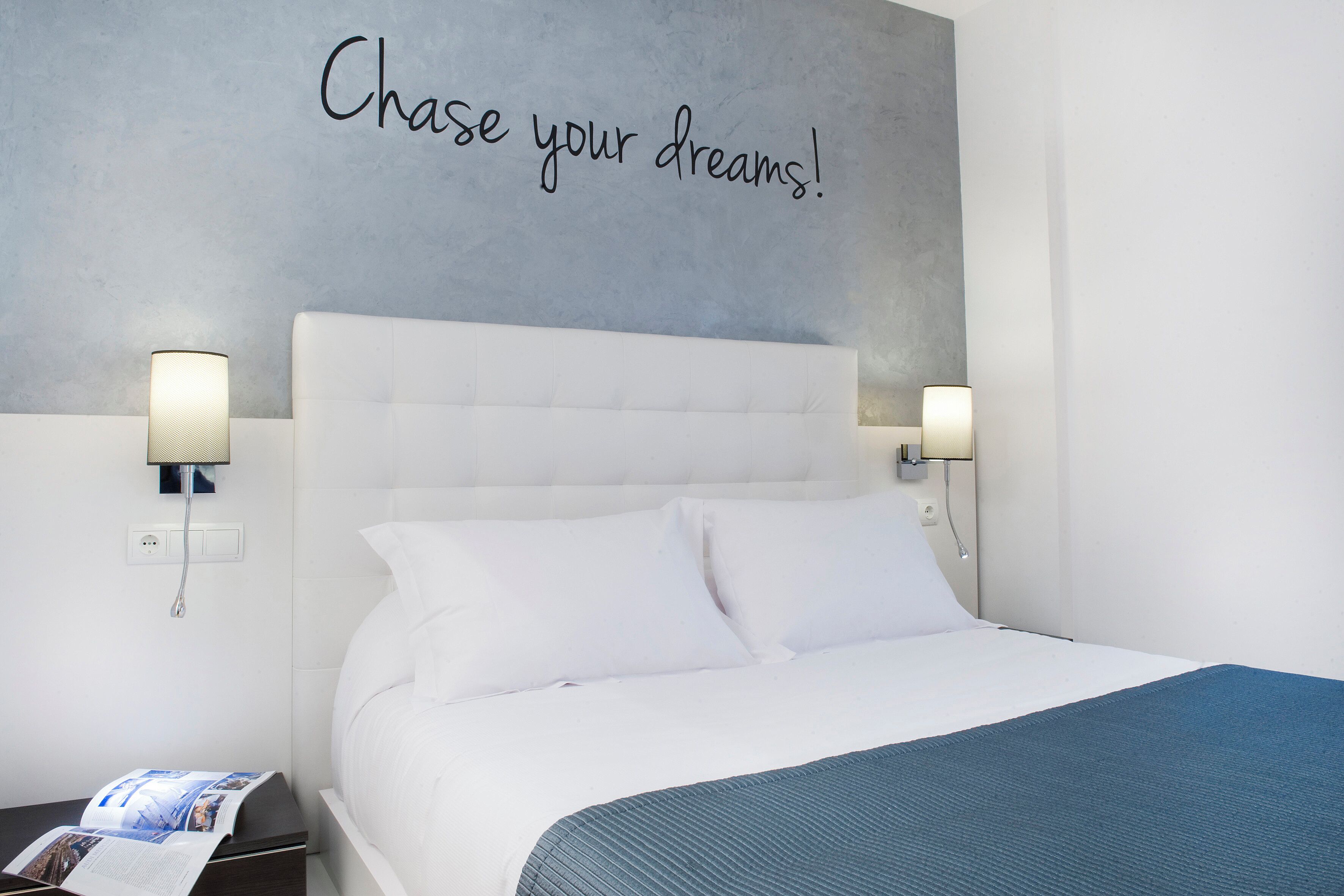 A neatly made bed with white linens and a blue throw blanket, two bedside lamps, and a motivational quote on the wall.
