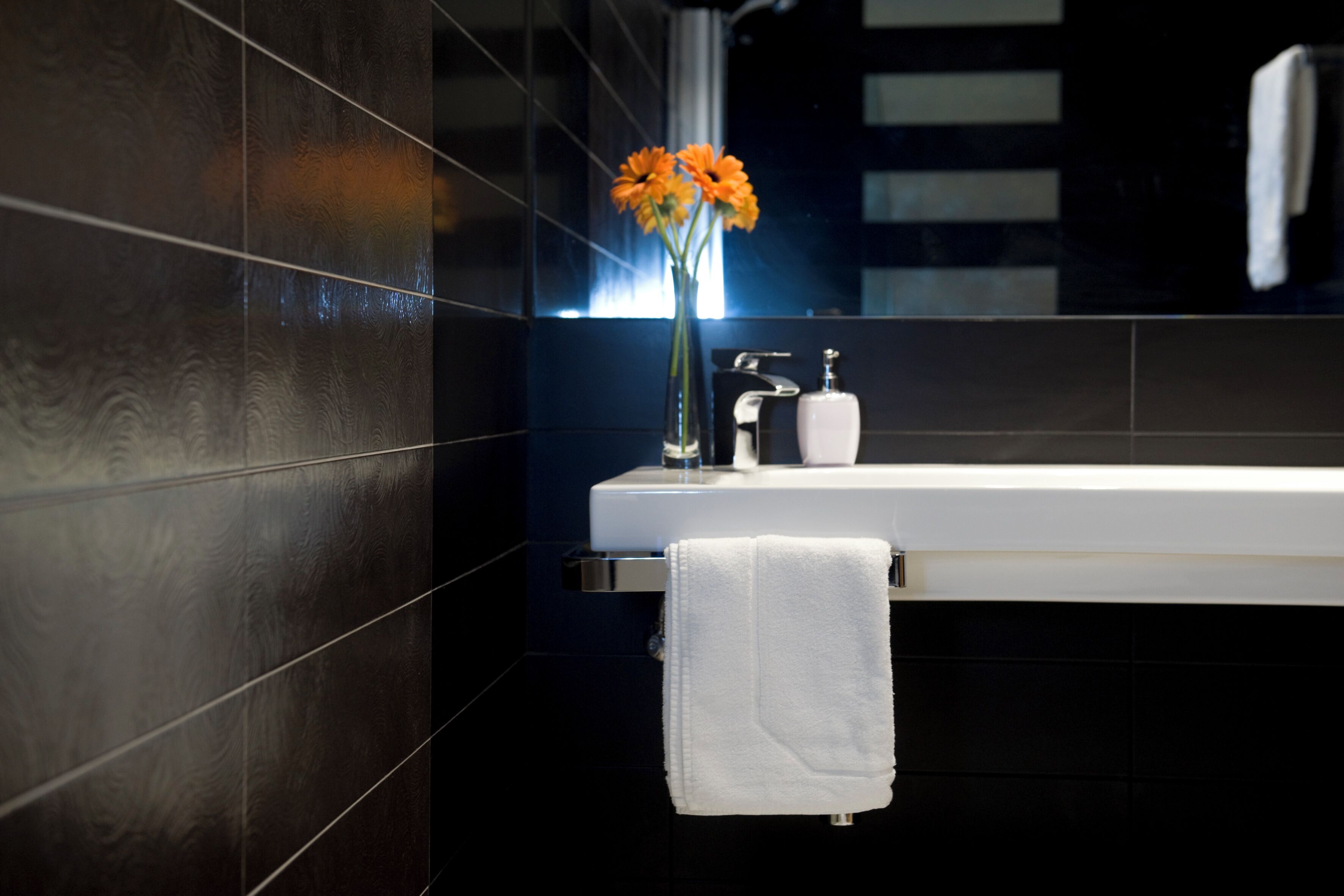 A modern bathroom with dark tiles, a white sink, a mirror, and a vase with orange flowers.