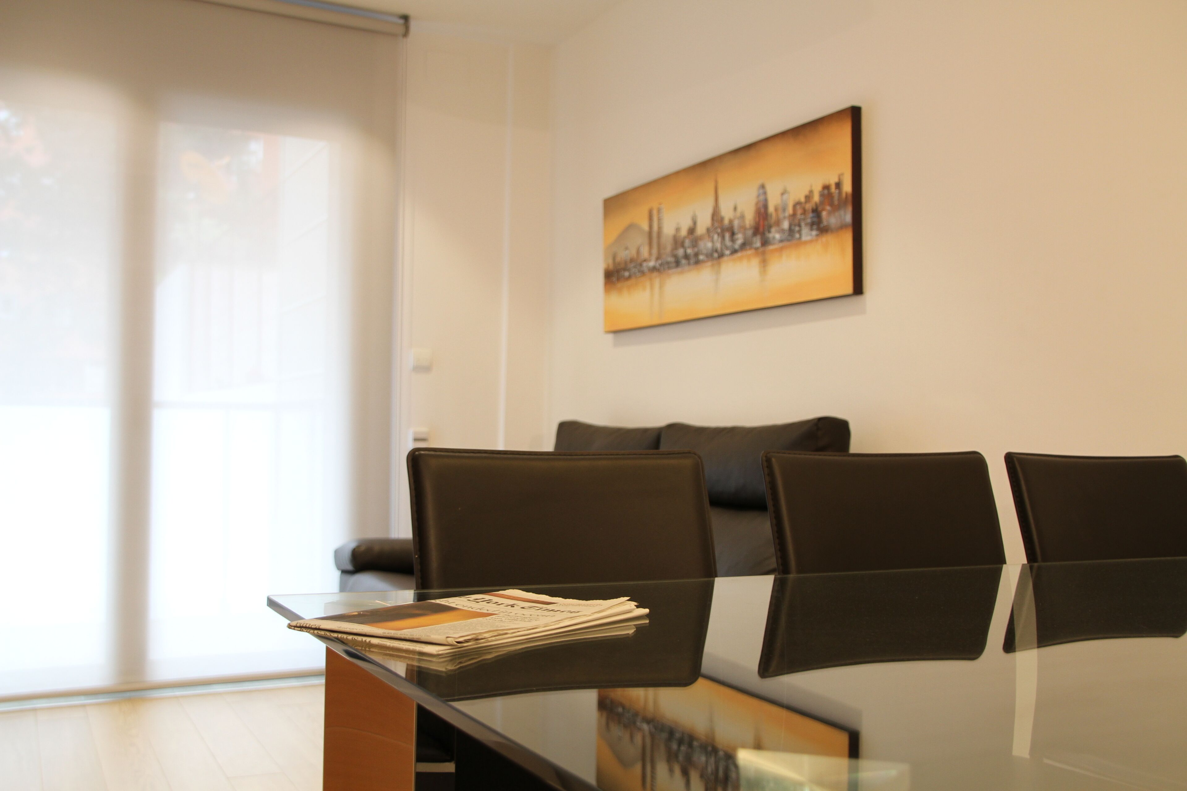 Een moderne kamer met een glazen tafel, zwarte stoelen, een bank en een stadslandschap schilderij aan de muur.