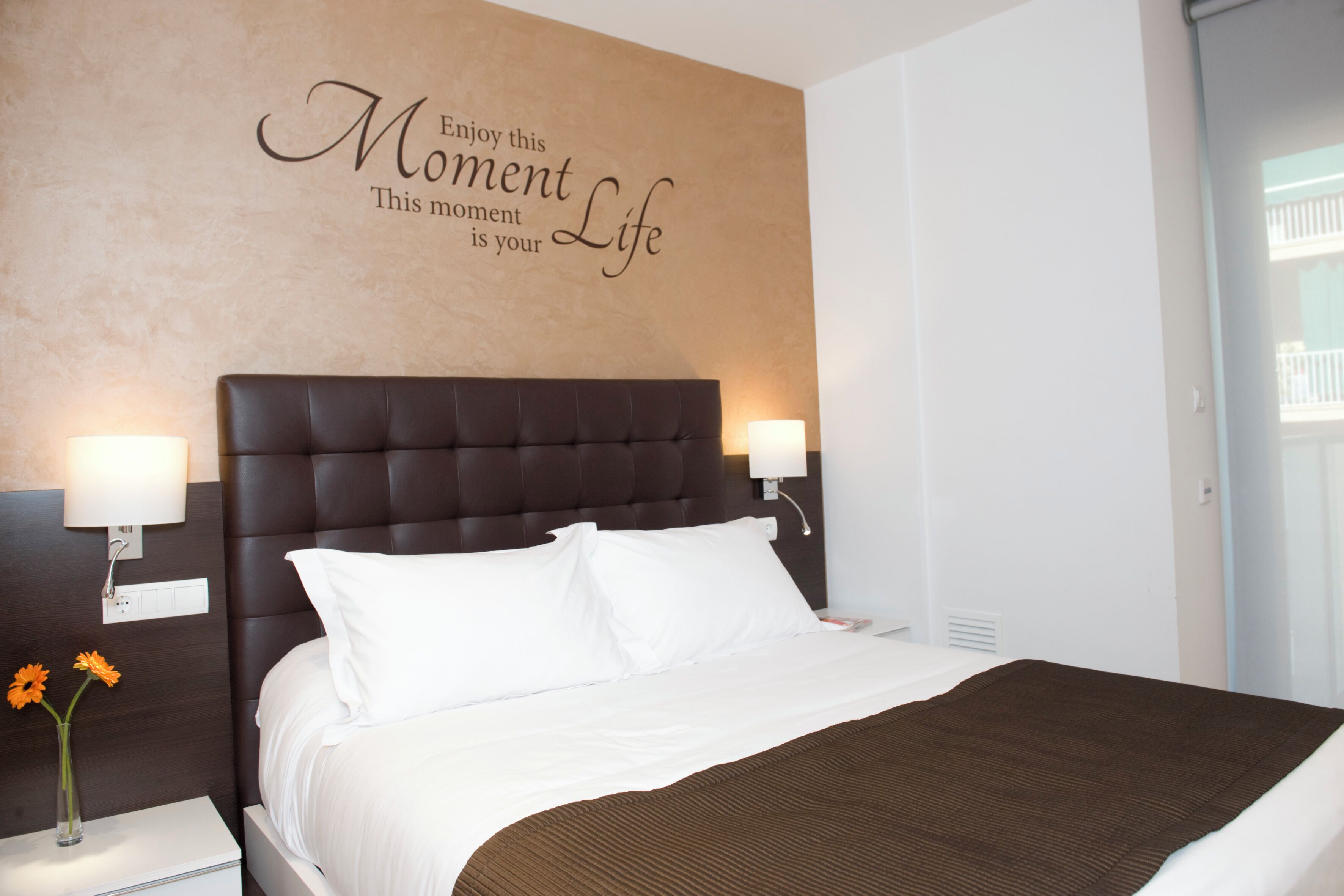 Habitación de hotel con una cama grande, dos lámparas de noche, una pared con una cita motivacional y una ventana con vista a un balcón.