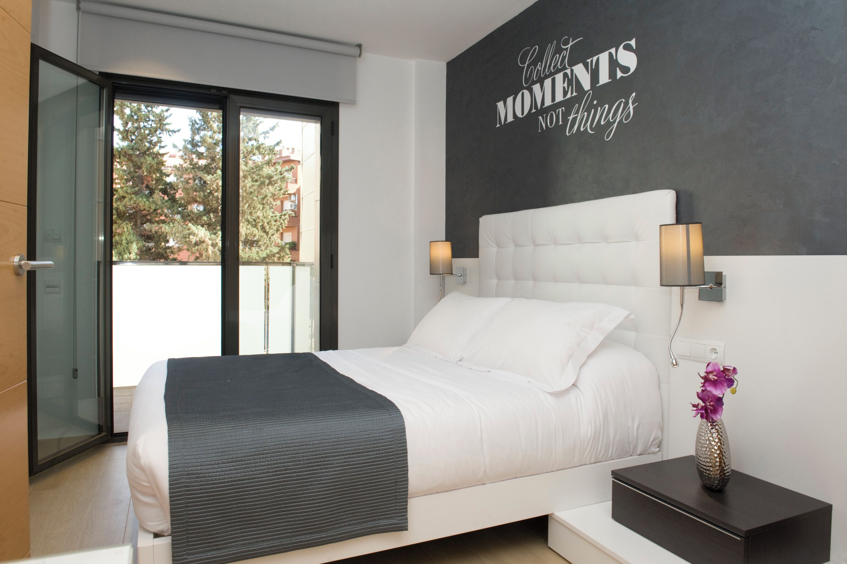 Een moderne slaapkamer met een groot bed, een nachtkastje met een vaas, en een muur met een motiverende quote.