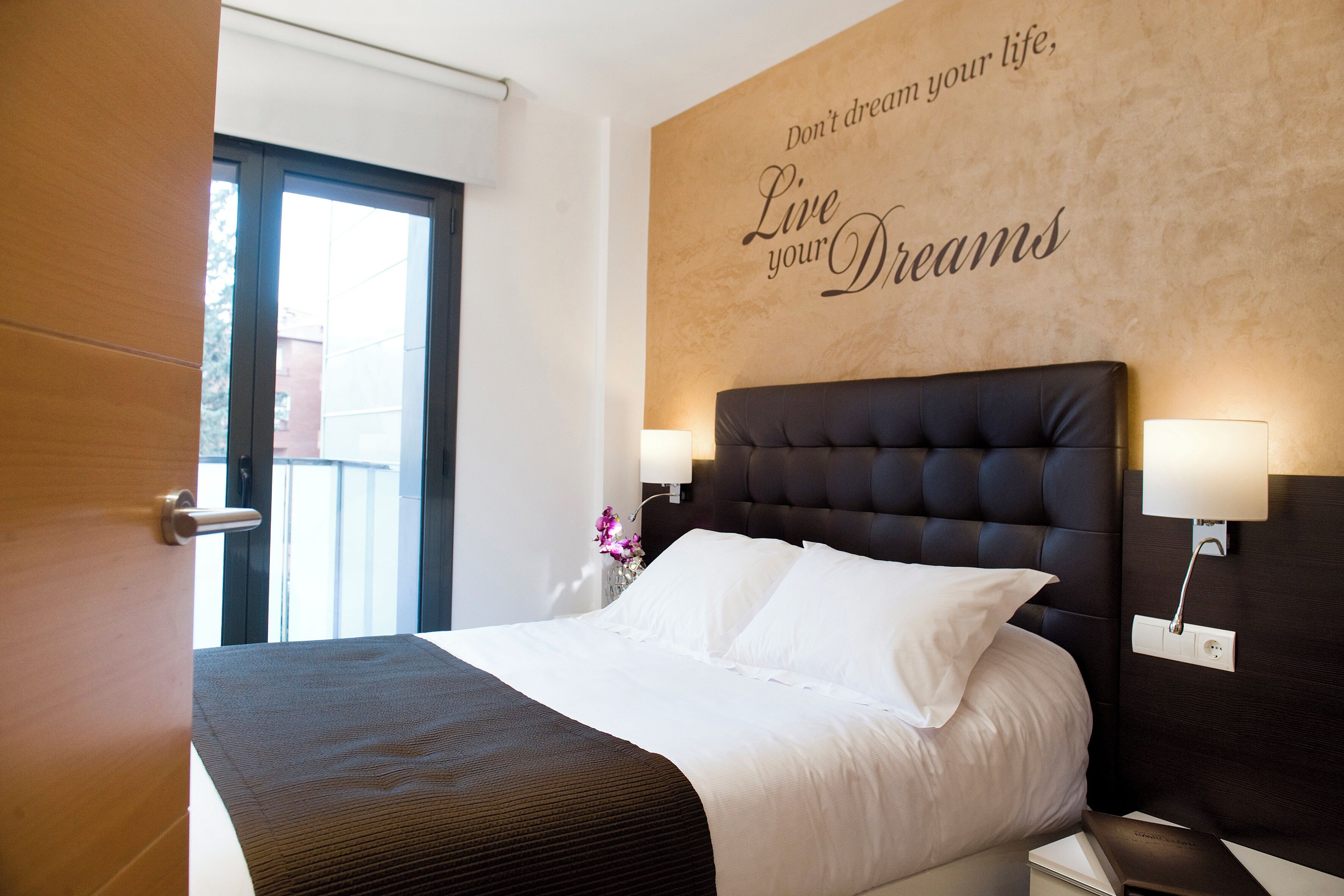 Habitación de hotel con una cama grande, una pared con texto motivacional, una mesita de noche con lámpara y vistas a través de una puerta.