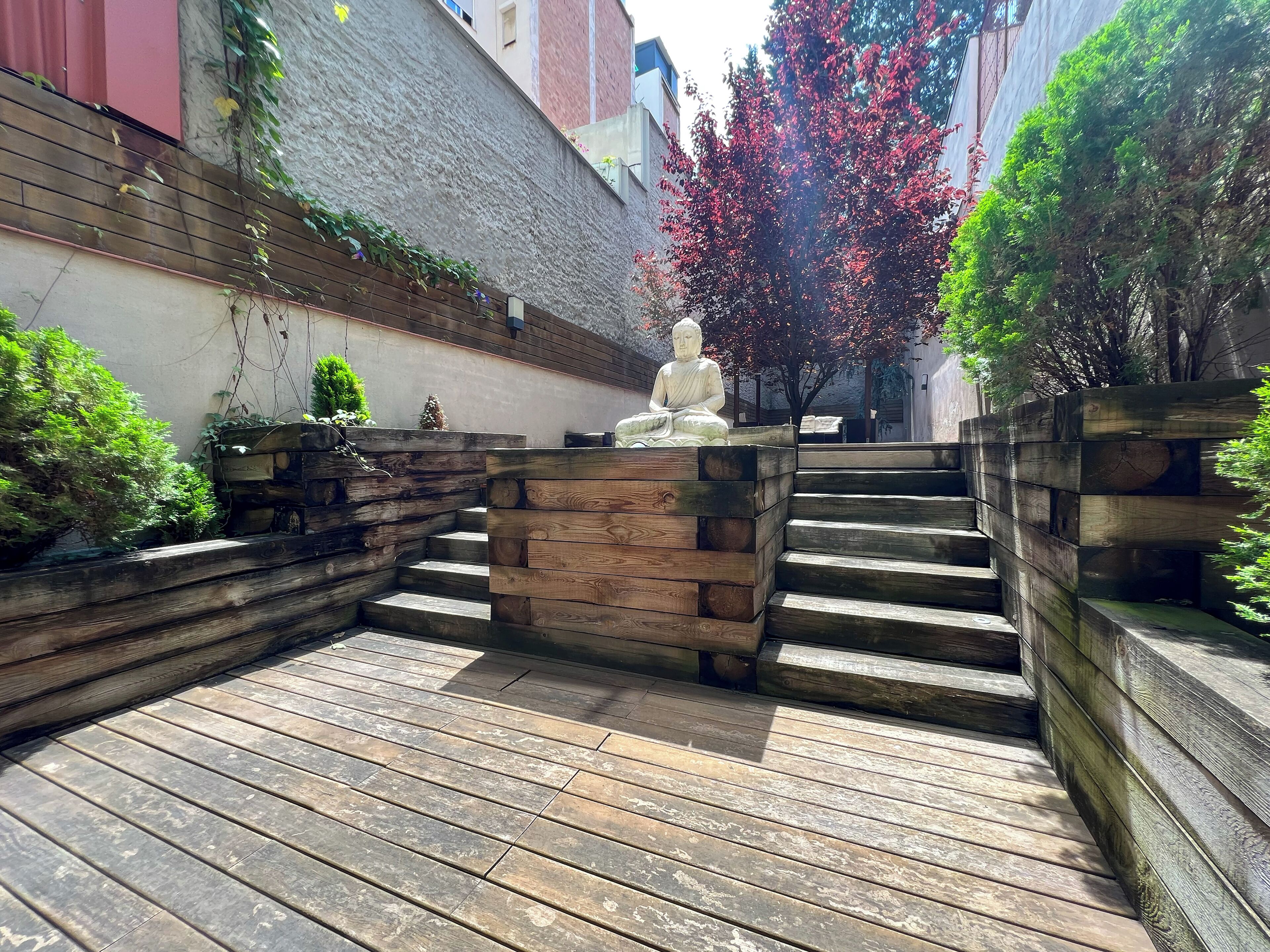 A wooden staircase leading to a seating area with a Buddha statue, surrounded by greenery and a tree with red leaves.