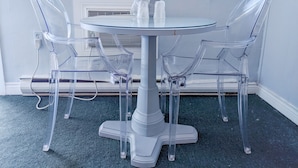 Una mesa redonda de vidrio con cuatro sillas transparentes, una base blanca tipo pedestal y una ventana con persianas al fondo.