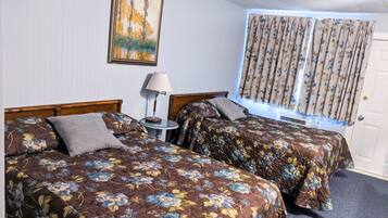 A hotel room with two beds, floral bedding, a bedside table with a lamp, a painting on the wall, and a window with curtains.