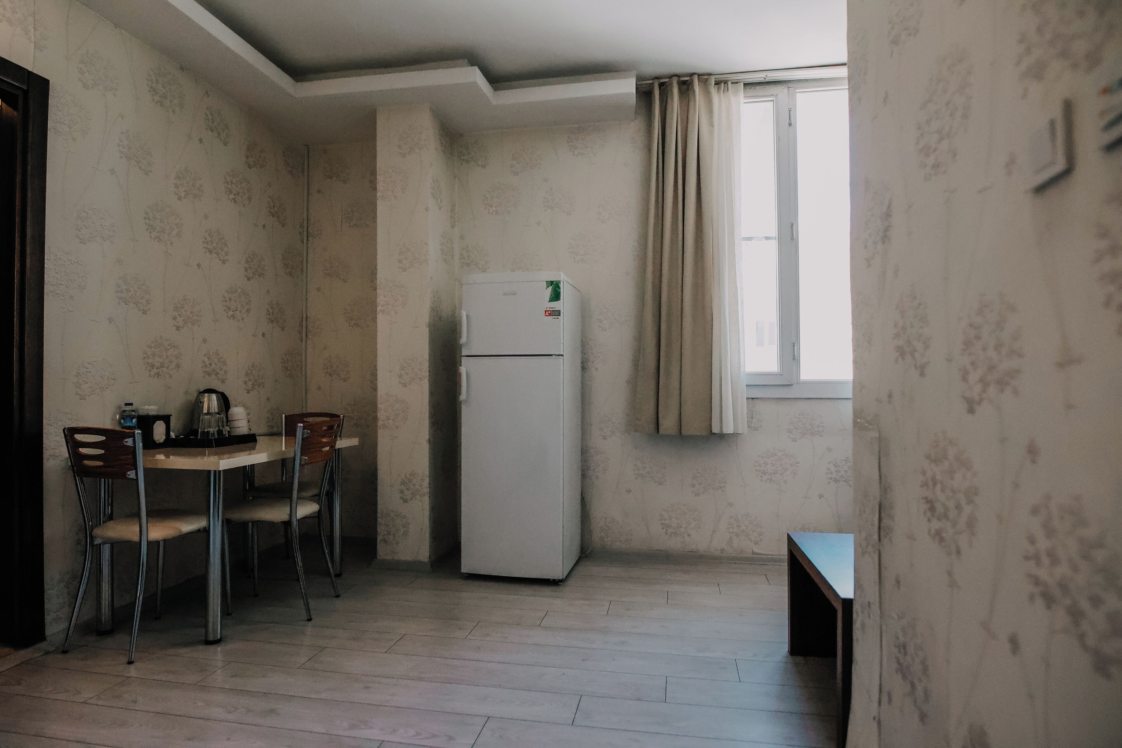 A small, sparsely furnished room with a single table and two chairs, a white refrigerator, and a window with curtains.