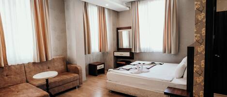 Una habitación de hotel moderna con sofá, una mesita redonda pequeña, una cama con colchón y almohadas blancas, una mesita de noche con lámpara y un espejo grande sobre la cama.