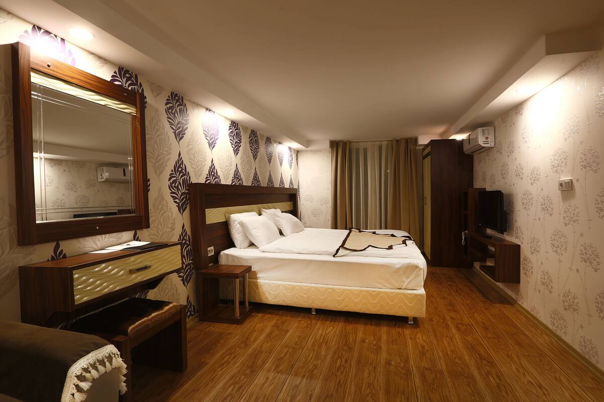 Hotellihuone, jossa on sänky, puinen lipasto, peili, televisio ja ikkuna, jossa on verhot.