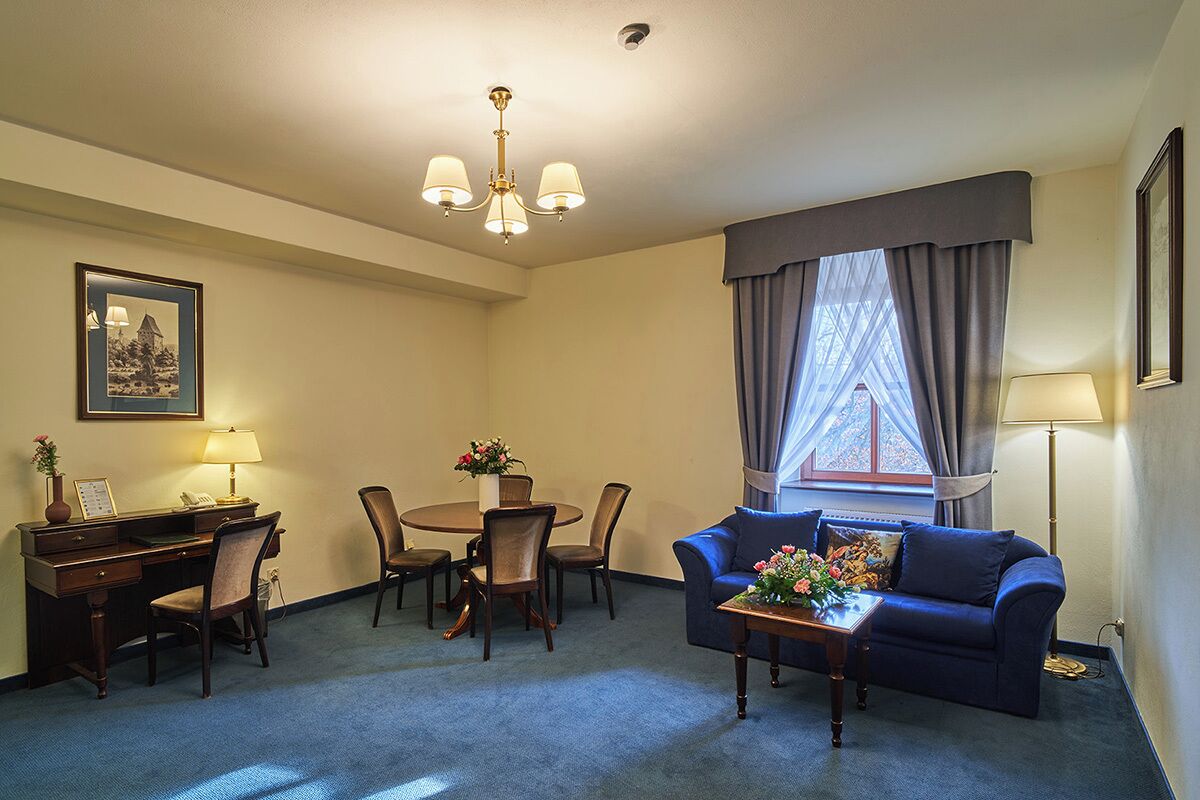Une chambre d’hôtel comprenant un coin repas, un canapé, un bureau avec une lampe et une fenêtre avec des rideaux.