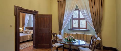 Un comedor con una mesa redonda, sillas y una lámpara de araña. Se ve una ventana con cortinas y vistas a árboles desde el exterior.