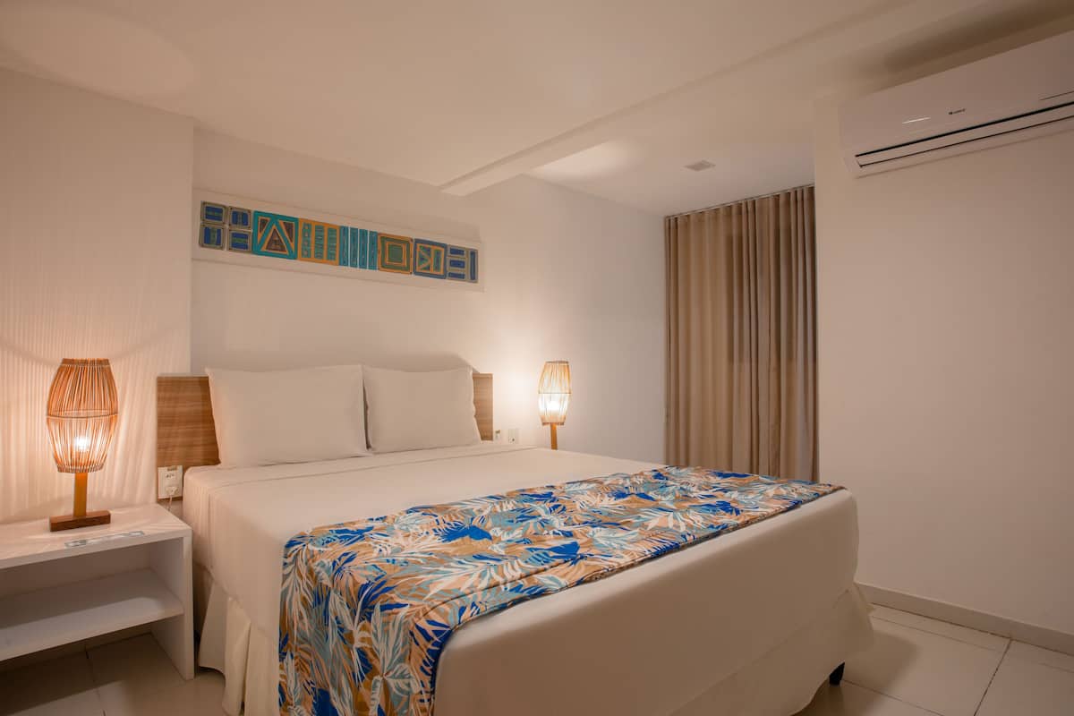A hotel room with a large bed, two bedside lamps, a wall-mounted artwork, and an air conditioning unit.