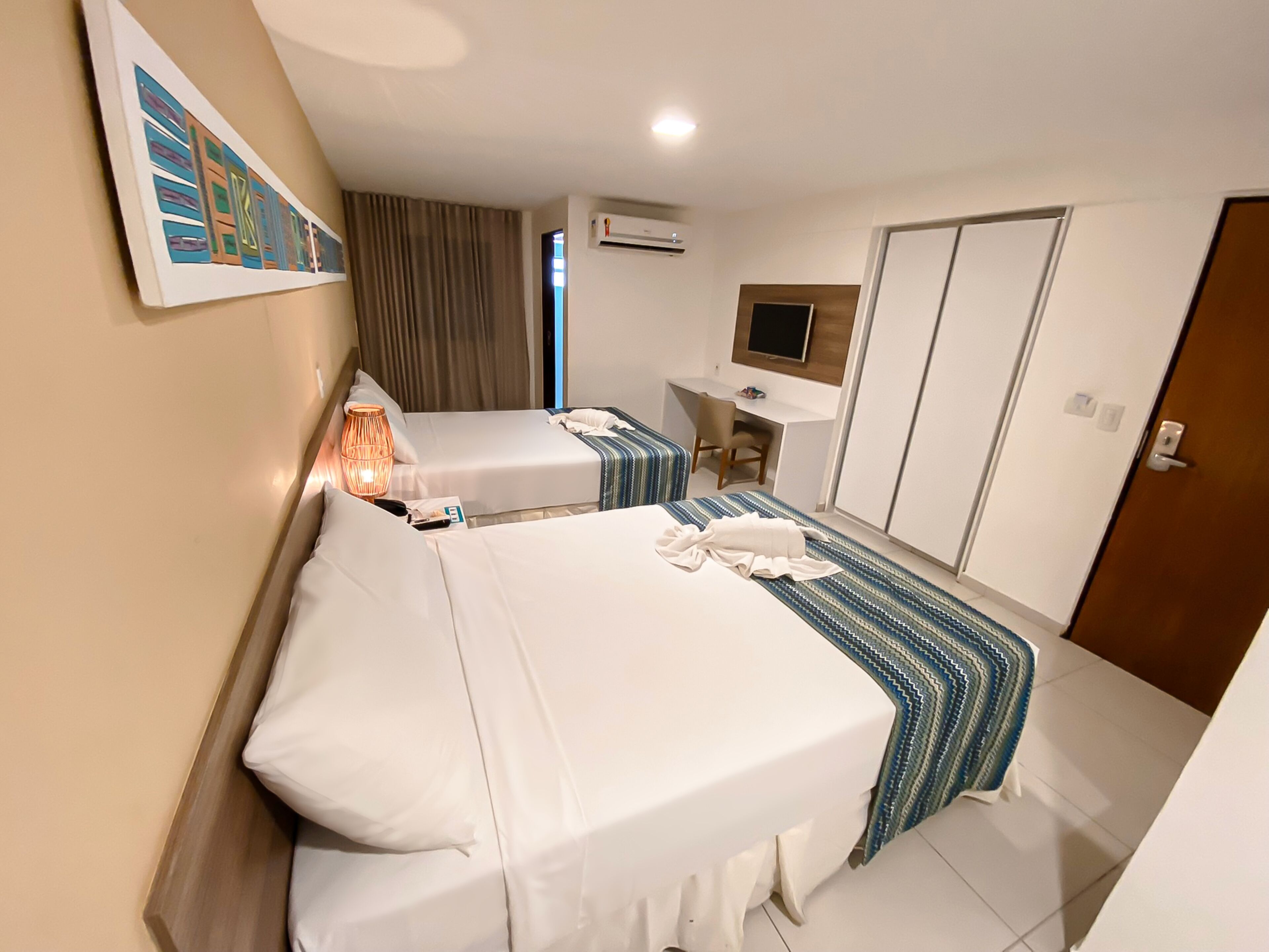A hotel room with two beds, a desk, a chair, a TV, and an air conditioning unit.