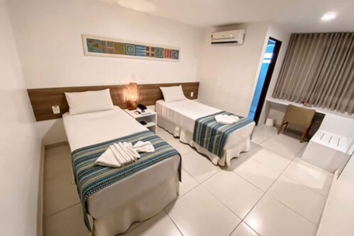 A hotel room with two beds, a desk, a chair, a lamp, and an air conditioning unit.