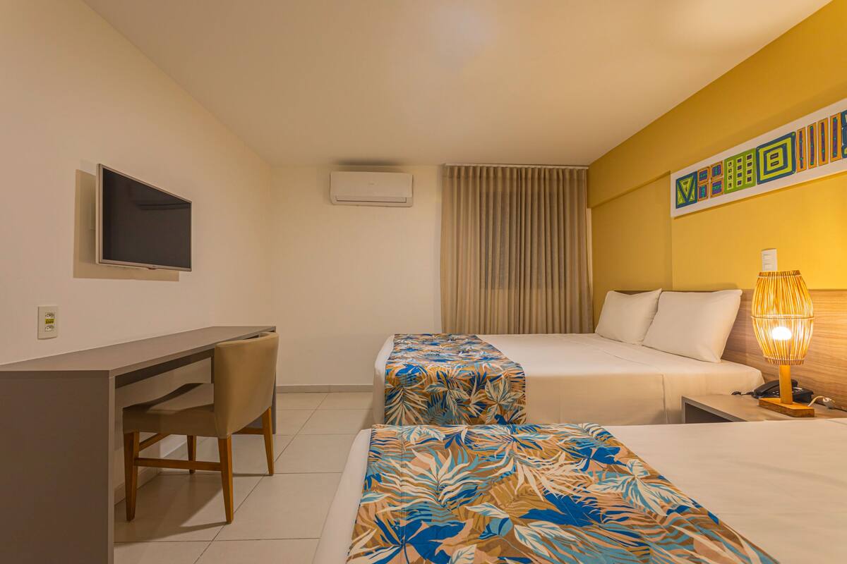 A hotel room with a desk, two beds, a TV, an air conditioner, a lamp, and a curtain.