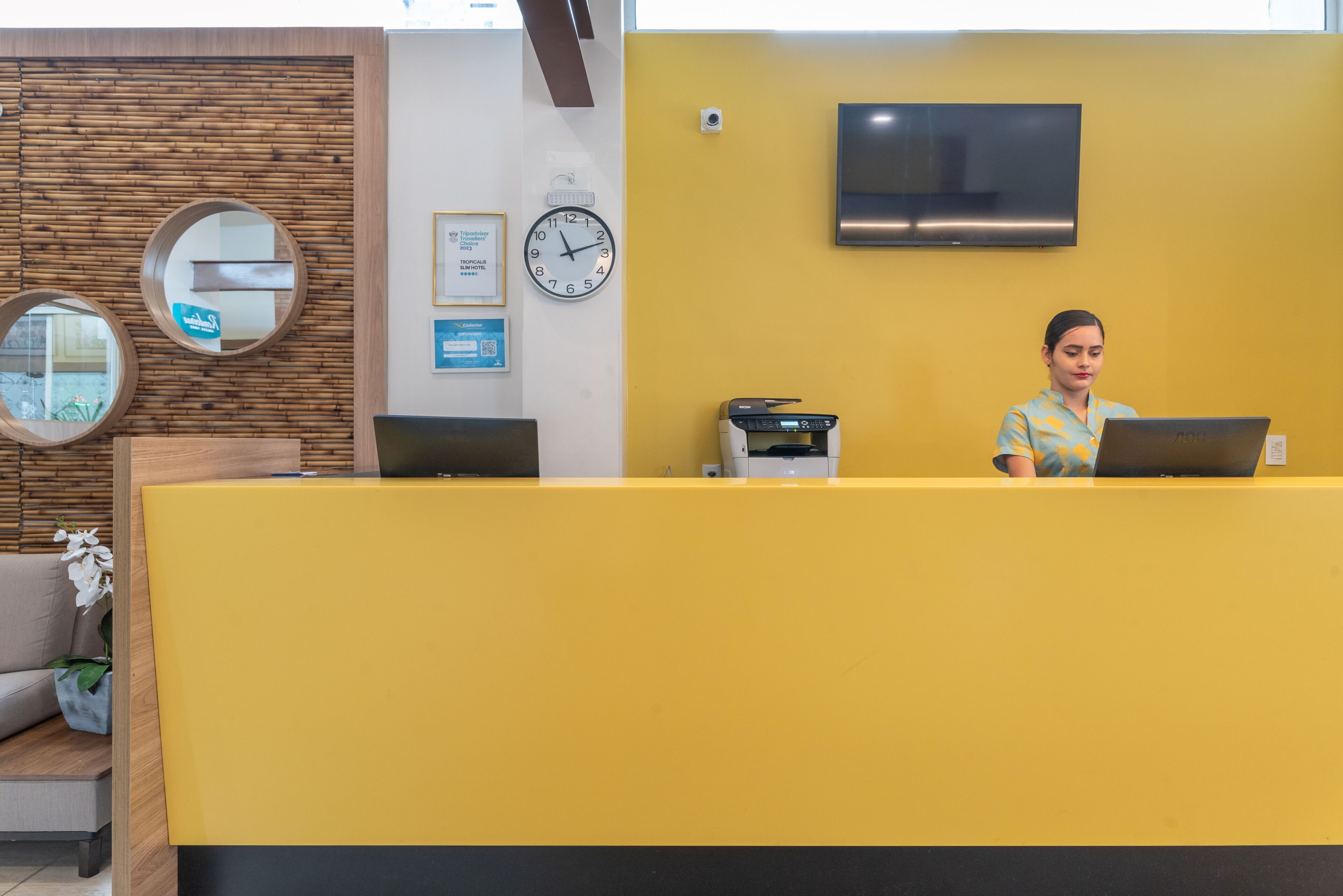 Uma área de recepção com balcão amarelo, um relógio e uma televisão na parede.
