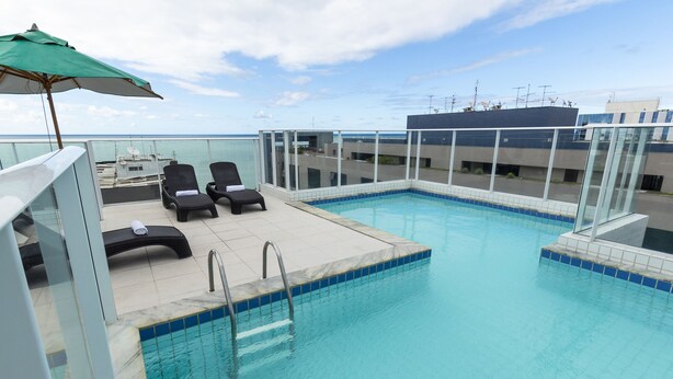 A rooftop pool with clear blue water, a glass balustrade, and two lounge chairs under a green umbrella. - Slim Pajuçara by Tropicalis (Maceió)