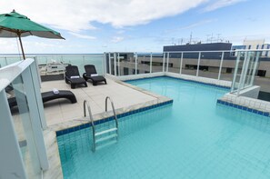 Une piscine sur le toit, avec une eau d’un bleu limpide, une balustrade en verre et deux chaises longues placées sous un parasol vert.