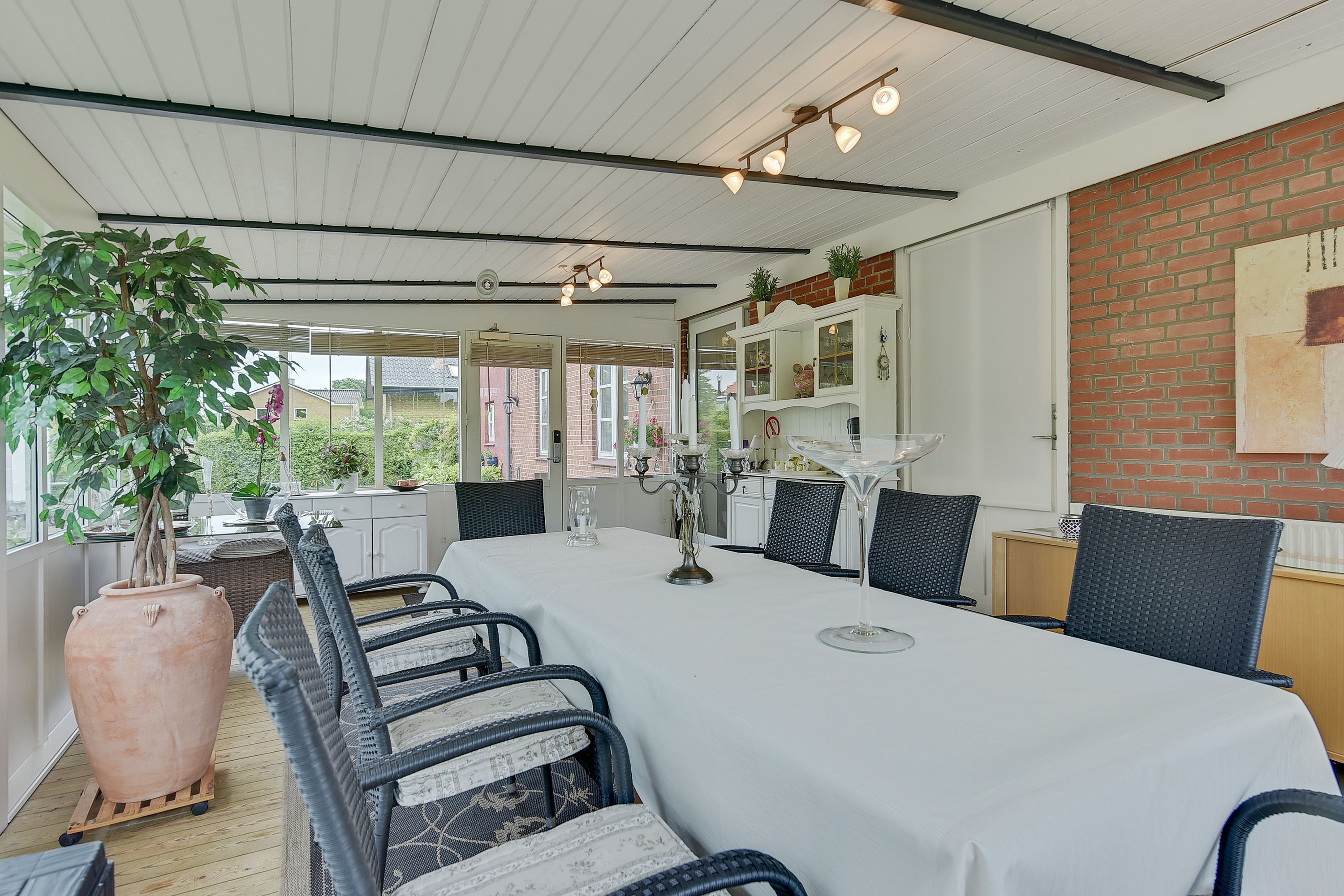 A dining area with a long table, wicker chairs, a potted plant, and a brick wall.