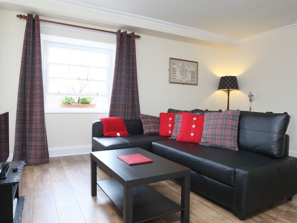 A modern living room with a black leather sofa, red cushions, a wooden coffee table, and a window with plaid curtains.