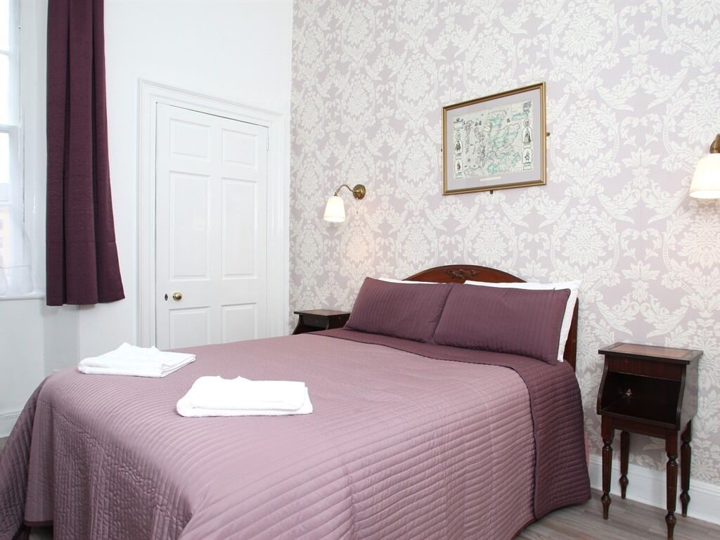 A neatly made bed with a purple bedspread, a white door, patterned wallpaper, and a framed picture on the wall.