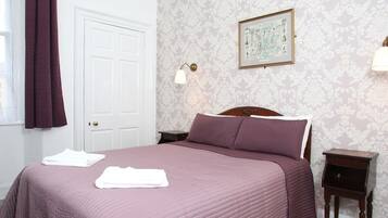 A neatly made bed with a purple bedspread, a white door, patterned wallpaper, and a framed picture on the wall.