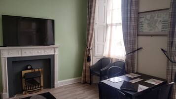 Een kamer met een open haard, een televisie erboven, een eettafel met stoelen en een raam met gordijnen.