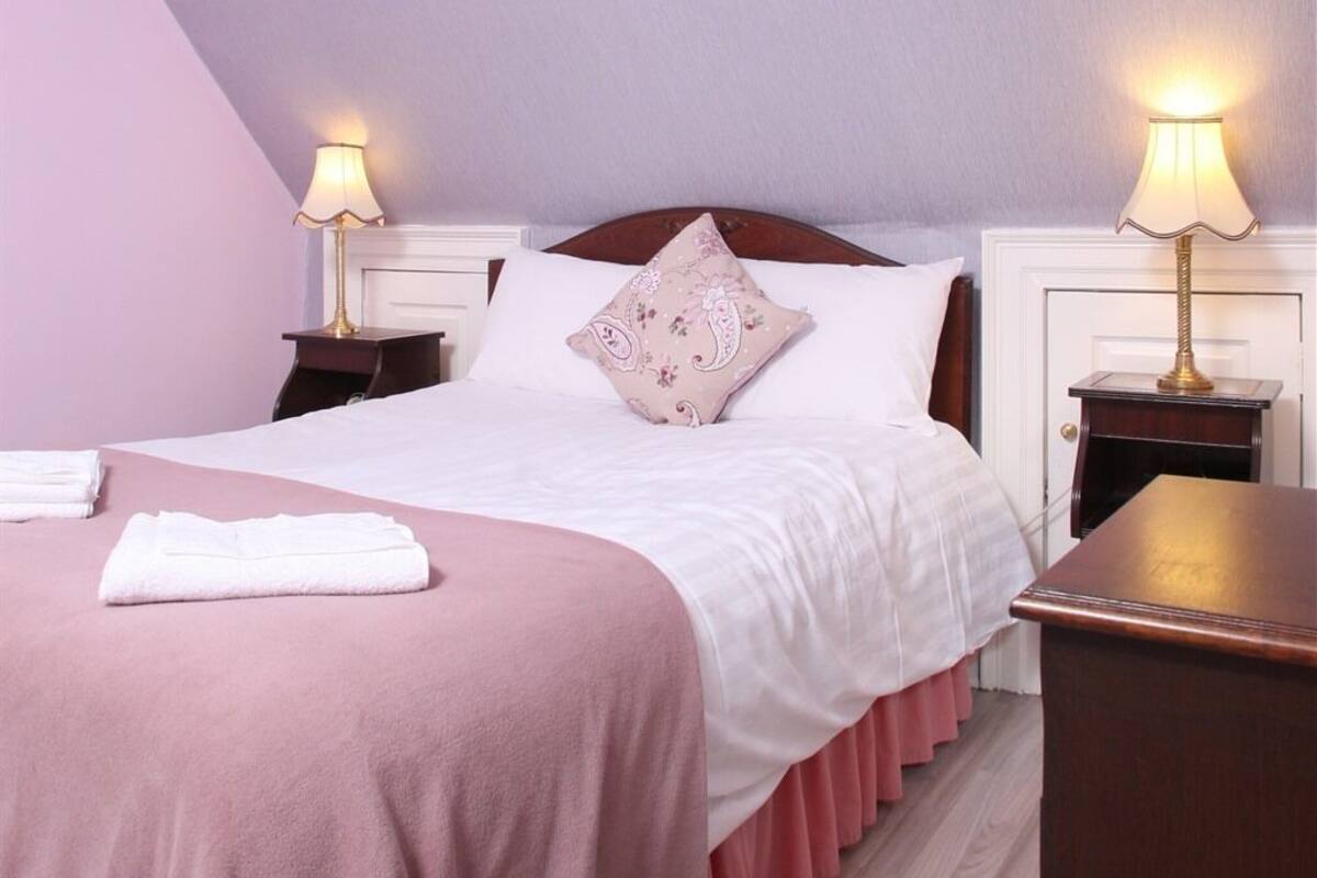 Ein ordentlich bezogenes Bett mit einer rosafarbenen Tagesdecke und weißen Kissen, zwei Nachttischlampen und einem Holznachttisch.