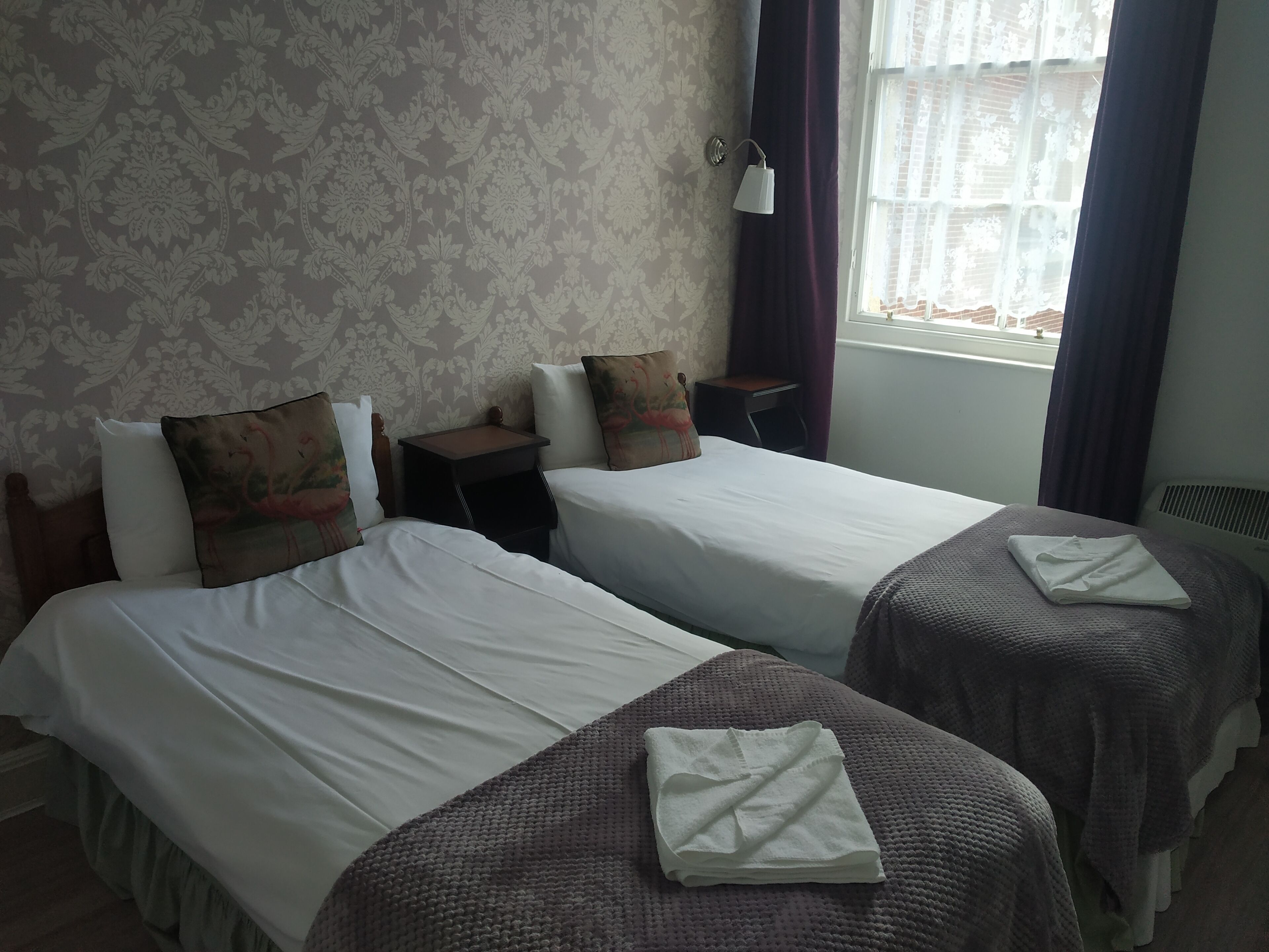 Habitación de hotel con dos camas, una mesita de noche, una lámpara y un ventanal con cortinas.