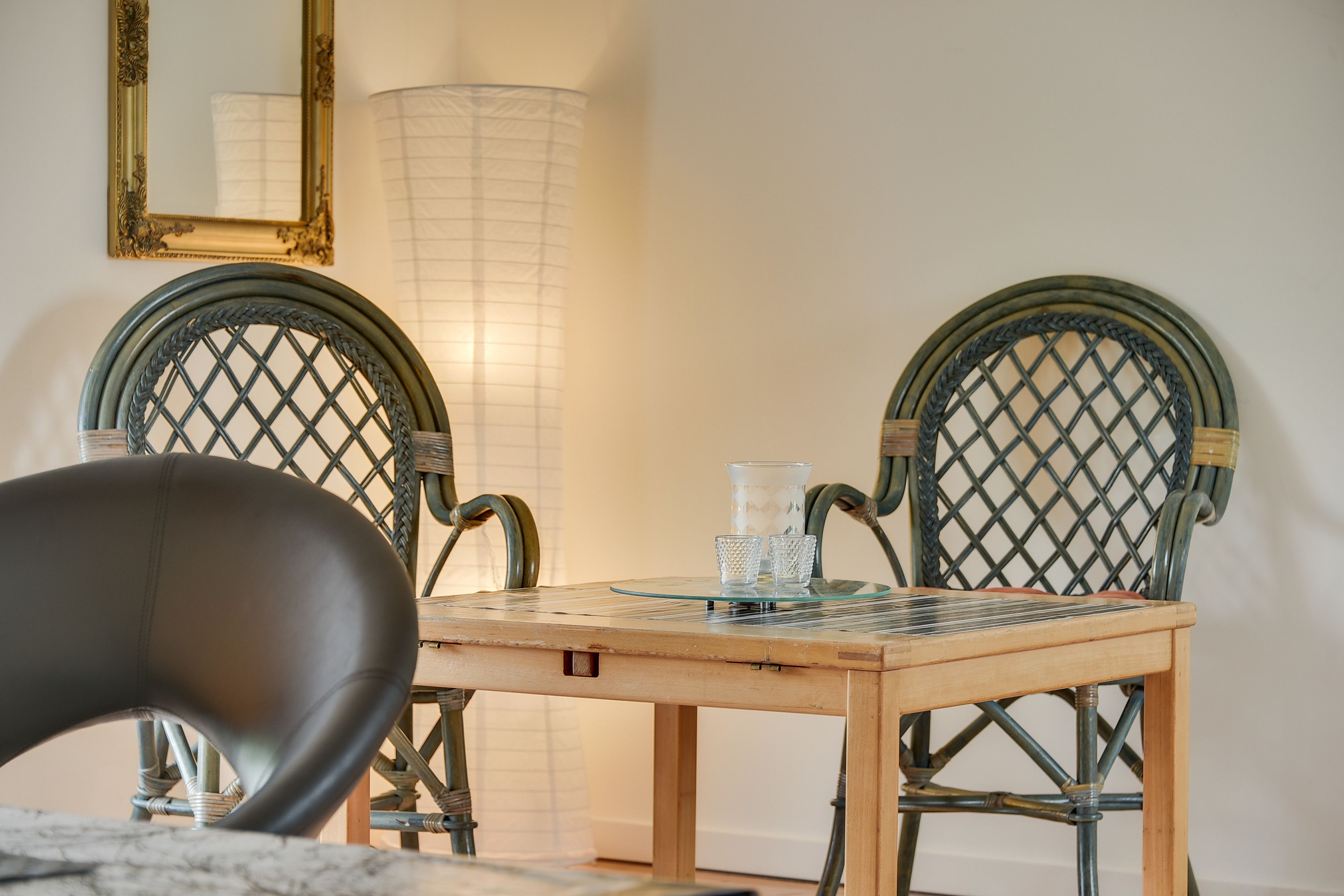A dining area with a wooden table, two wicker chairs, a glass candle holder, and a tall floor lamp.