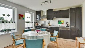 A modern kitchen with dark cabinets, a stainless steel range hood, and a dining area with a round table and teal chairs. - Bjerndrup Apartments (Aabenraa)