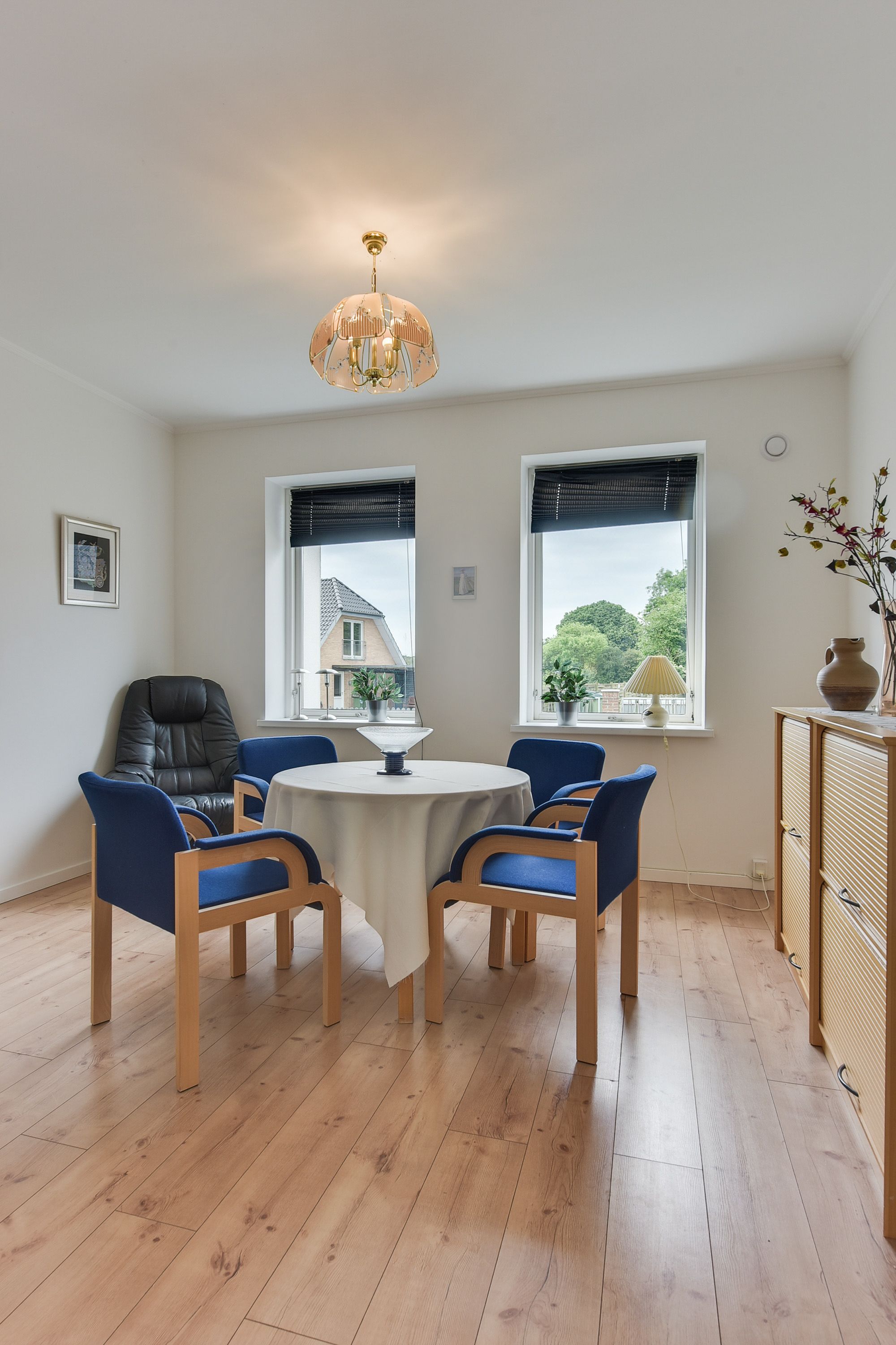 Un coin repas avec une table ronde, quatre chaises bleues, un fauteuil en cuir noir et un lustre. Il y a deux fenêtres équipées de stores, dont l’une donne sur une maison et l’autre sur des arbres.