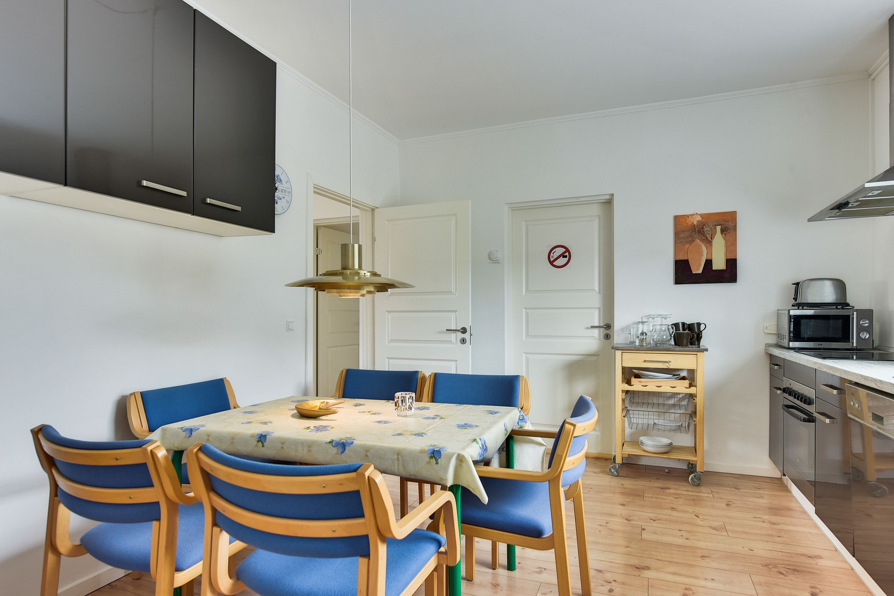 A dining area with a table set for six, surrounded by blue chairs. A kitchen with modern appliances is visible in the background.
