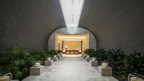 Um lobby com um balcão de recepção central, teto alto com luminárias pendentes e plantas em vasos dispostas simetricamente de ambos os lados.