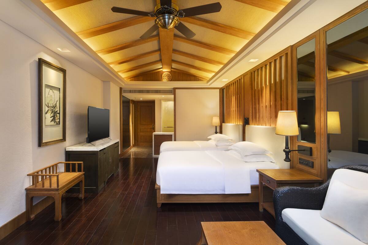 Habitación de hotel con techo de madera, una cama grande, dos mesitas de noche con lámparas, un televisor, un banco y un sofá.