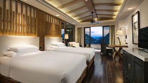 Quarto de hotel com duas camas, uma escrivaninha, uma cadeira, uma janela grande com vista para as montanhas e um ventilador de teto.