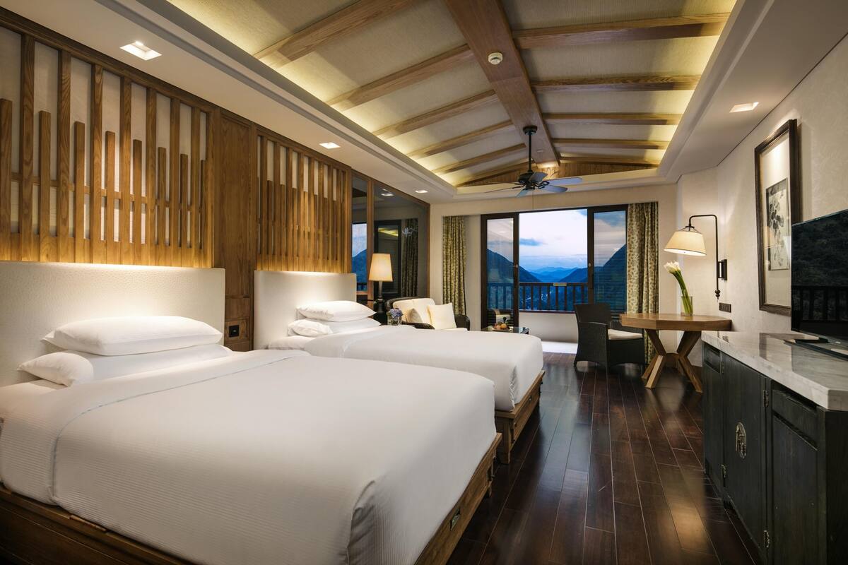 Habitación de hotel con dos camas, un escritorio, una silla, un ventanal con vistas a la montaña y un ventilador de techo.