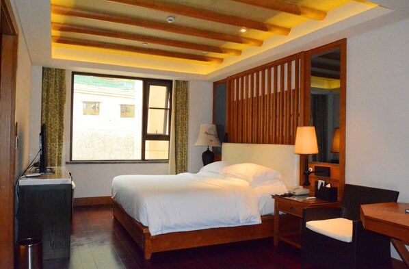 Habitación de hotel con cama de madera, un escritorio, una silla, una lámpara y un ventanal grande.